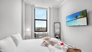 A hotel room with a large bed, a TV mounted on the wall, a window with a city view, and a desk with a lamp. - XL Studio - Modern Studio with Rooftop Pool (Philadelphia)