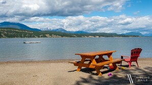 Escena de pícnic al costat d'un llac, amb una taula de fusta i una cadira vermella, una barca petita a l'aigua i muntanyes al fons.