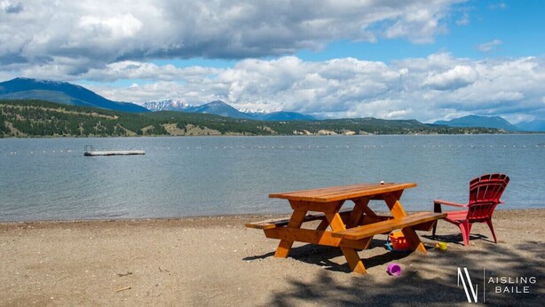A lakeside picnic setup with a wooden table and a red chair, a small boat on the water, and mountains in the background.