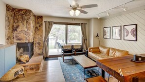 A living room with a stone fireplace, a sofa, a coffee table, and a dining table. There is a ceiling fan, a window with curtains, and framed artwork on the wall.