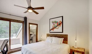 A bedroom with a bed, a ceiling fan, a wooden headboard, a nightstand, a lamp, a chair, a closet, and a balcony with a view of trees. - Cottage Culture | Indoor Pool & Hot Tub | Beach (Windermere)