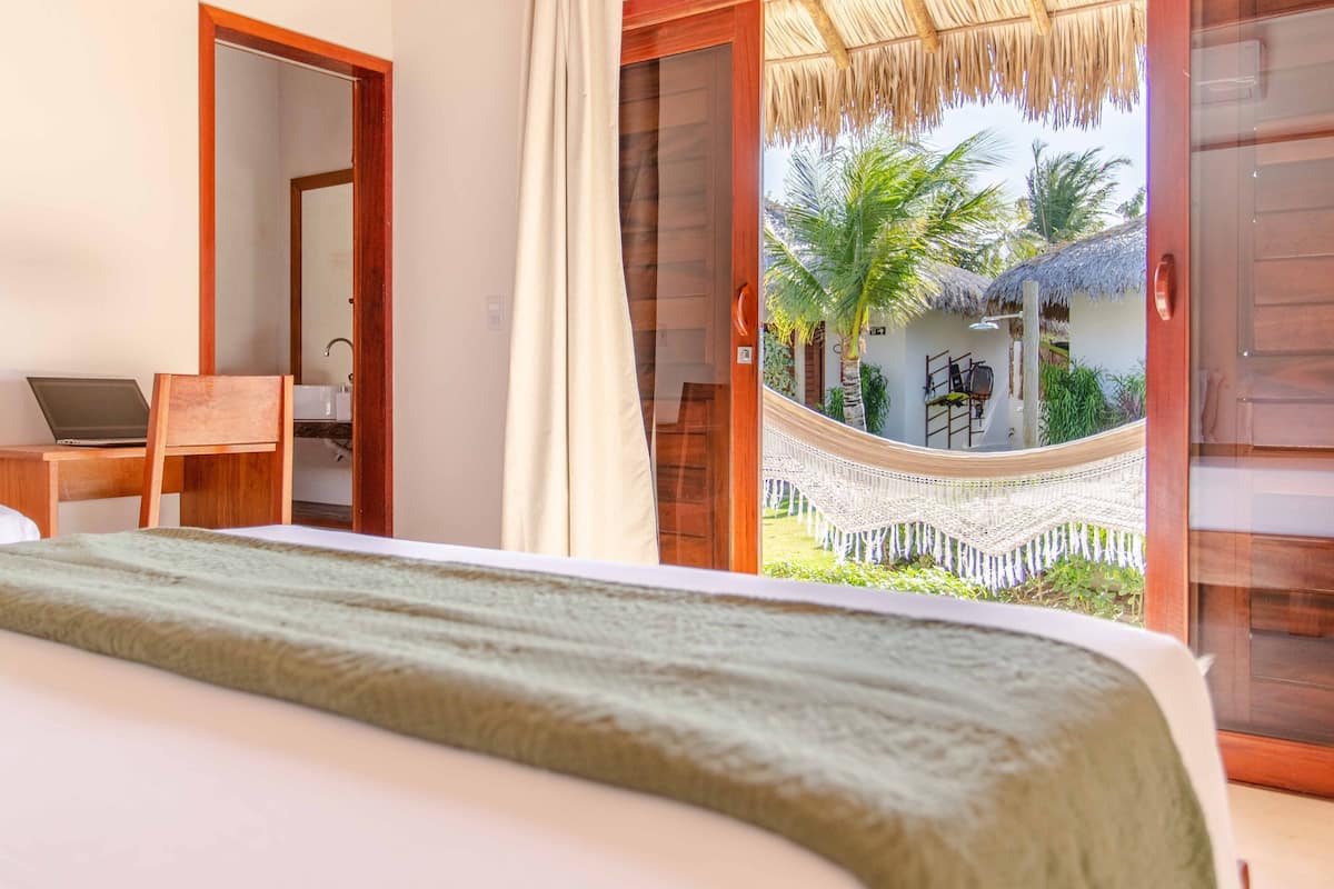 Un dormitorio con cama, una laptop sobre un escritorio y vistas al exterior, donde se ve una hamaca y palmeras.