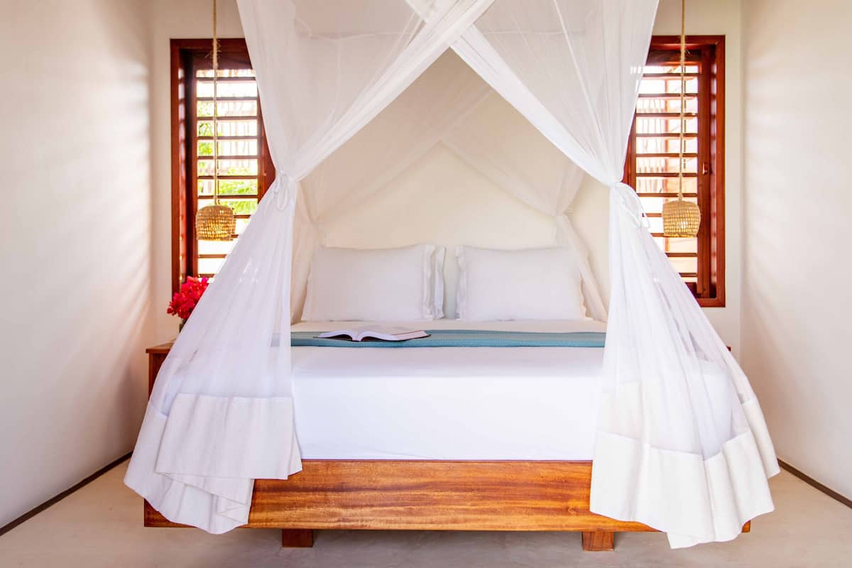 Una cama con dosel blanco, un banco de madera y una ventana con vistas a follaje.