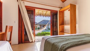 A bedroom with a bed, a wooden wardrobe, and a view of a thatched-roof house through a sliding door. - Vilarejo Preá (Cruz)