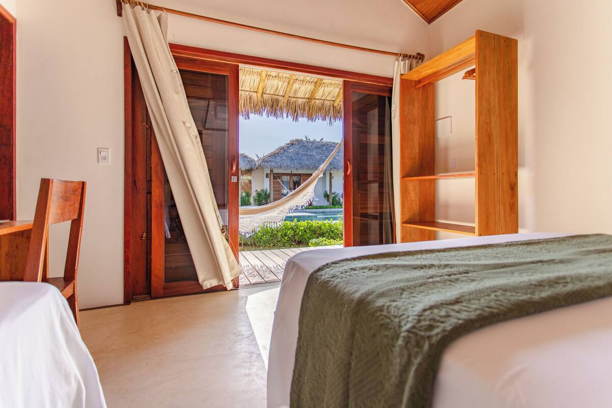 Un dormitorio con cama, un armario de madera y vista a una casa con techo de paja a través de una puerta corrediza.