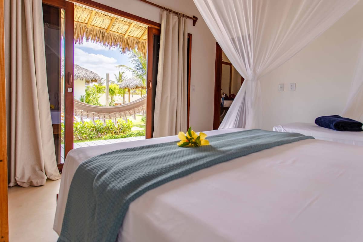Habitación de hotel con una cama, vista a una construcción de techo de paja y un balcón con vistas a la vegetación.