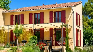 Une maison à deux étages avec un patio couvert, des volets rouges et un jardin où poussent différentes plantes et fleurs.