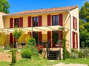 A two-story house with a covered patio, red shutters, and a garden with various plants and flowers. - Villa on Lac du Salagou (Octon)