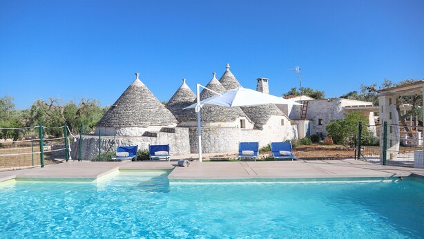 A swimming pool with blue water, surrounded by stone buildings with conical roofs, and blue lounge chairs. - TD Trulli Albapetra Pool & Olive Trees (Polignano a Mare)
