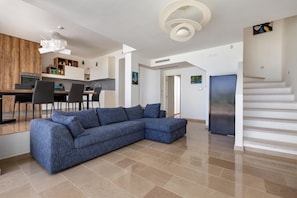 A modern living room with a sectional sofa, a dining area with a table and chairs, a kitchen with wooden cabinets, and a staircase.