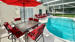 Zona de piscina con un patio de cristal, sillas rojas, una mesa y un paraguas rojo.