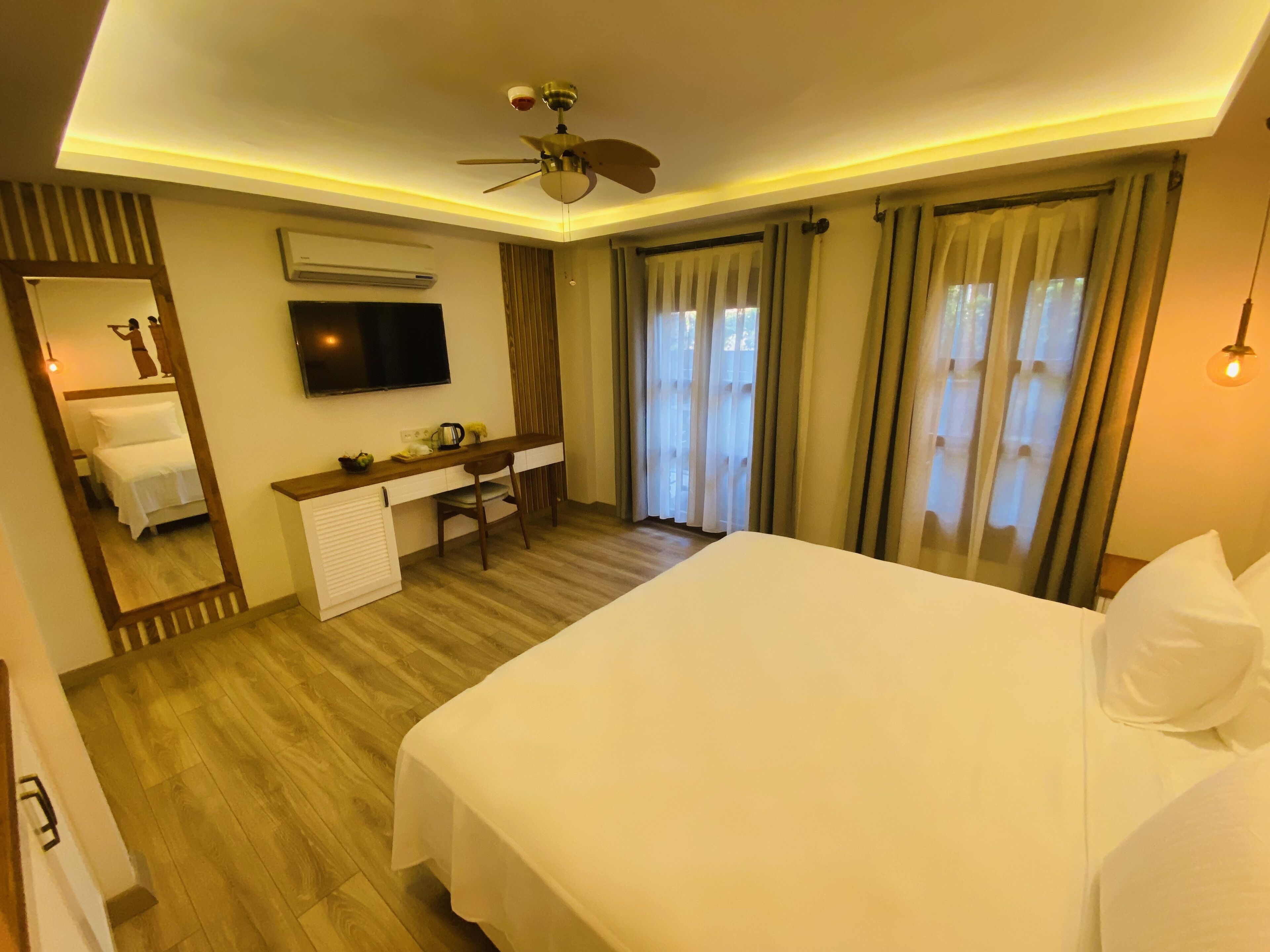 Habitación de hotel con una cama grande, un escritorio con silla, ventilador de techo, televisor de pantalla plana y un espejo amplio.