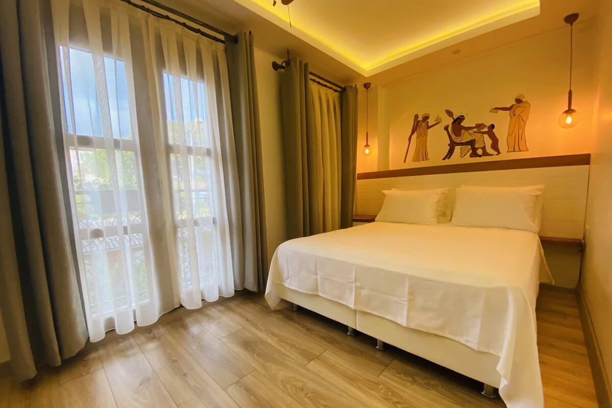 A hotel room with a large bed, wooden flooring, a balcony with curtains, and a wall with a mural.