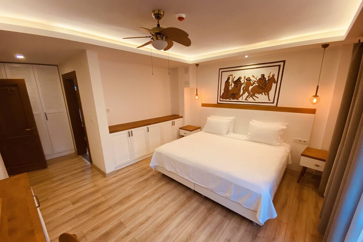 A bedroom with a bed, a ceiling fan, a nightstand, a wall-mounted light, and a door.