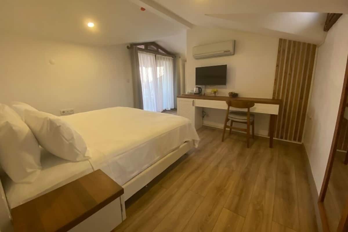 A bedroom with a bed, a desk, and a chair. There is a window with curtains, an air conditioning unit, and a television mounted on the wall.