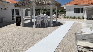 Un espace extérieur aménagé avec des sièges, abrité par un toit de chaume, entouré de chaises blanches et d’une piscine.