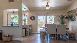 A spacious living area with a ceiling fan, a dining table with chairs, a kitchen area with a counter, and a large window with a view of palm trees.
