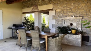 Eine Terrasse im Freien mit einer Steinmauer, einem Holztisch, Korbstühlen, einem Grill und Topfpflanzen.