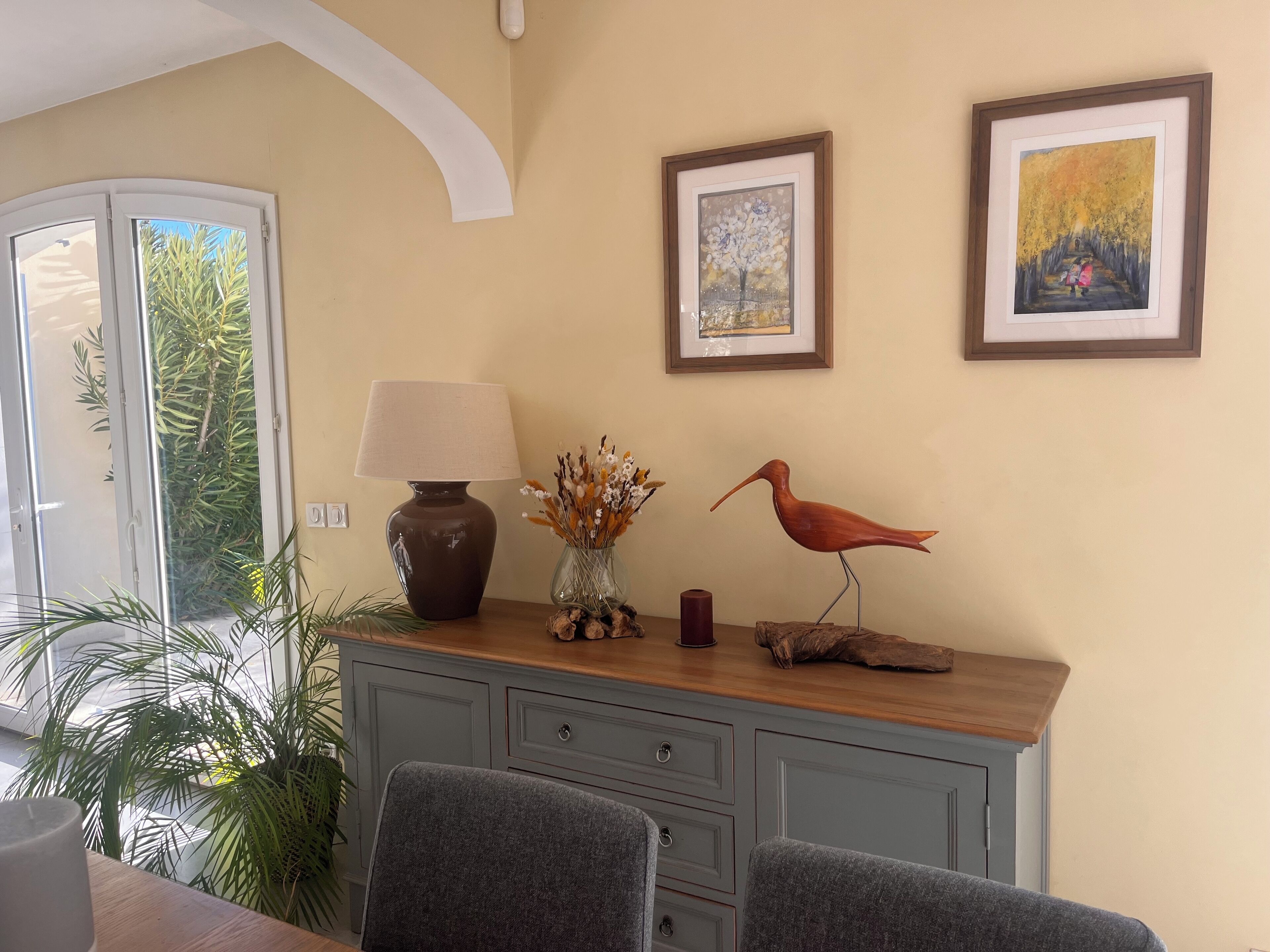 A dining area with a table, chairs, a lamp, a vase with dried flowers, and two framed artworks on the wall.