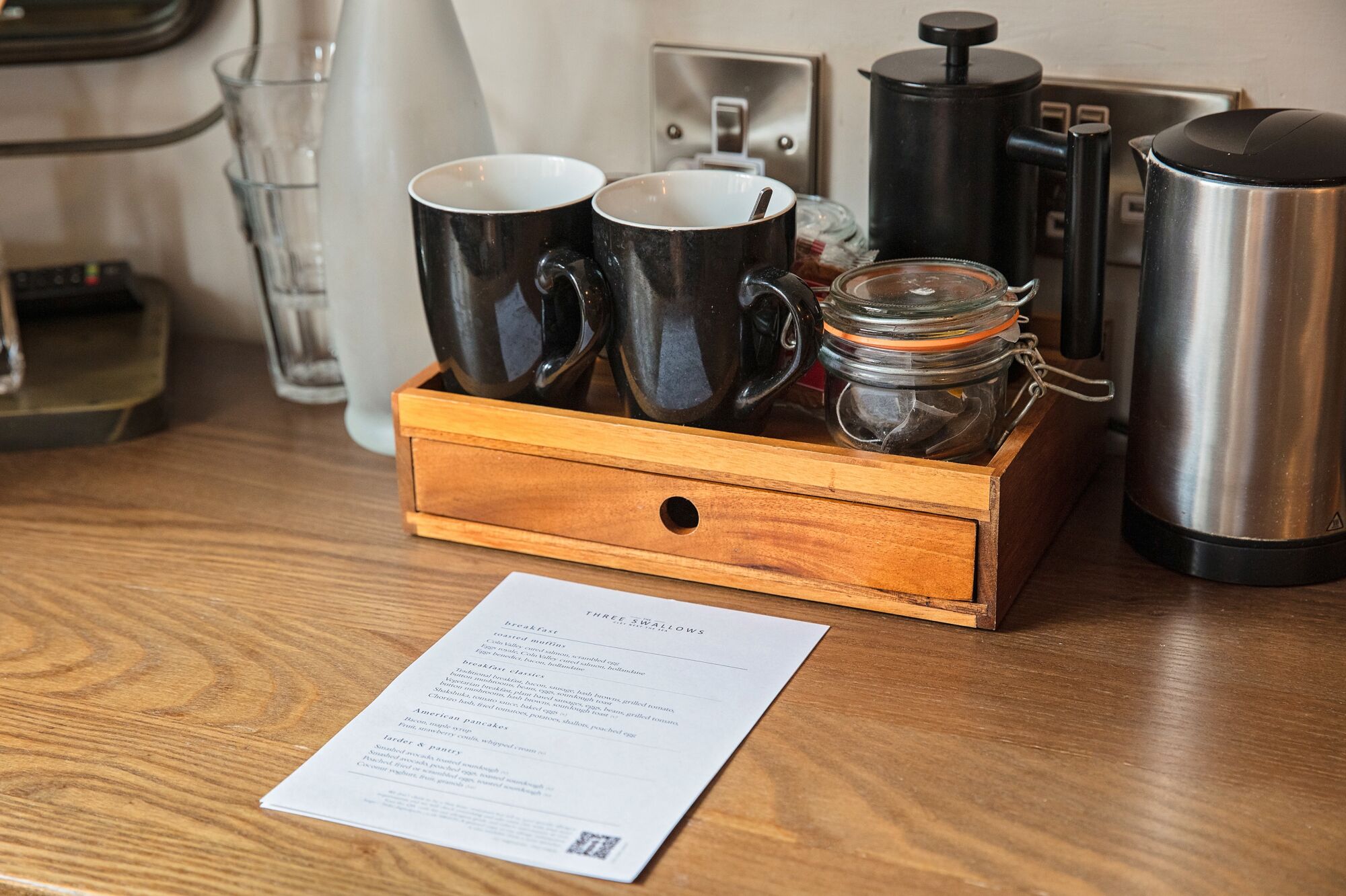 A wooden tray with two black mugs, a glass jar, and a stainless steel coffee pot on a wooden table.