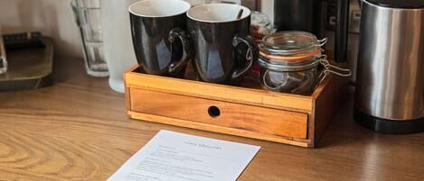 A wooden tray with two black mugs, a glass jar, and a stainless steel coffee pot on a wooden table.