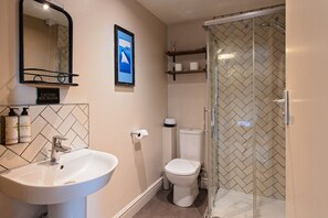 Un baño con lavamanos, inodoro y ducha. Hay un letrero de precaución por agua caliente, un cuadro en la pared y artículos de aseo en un estante.
