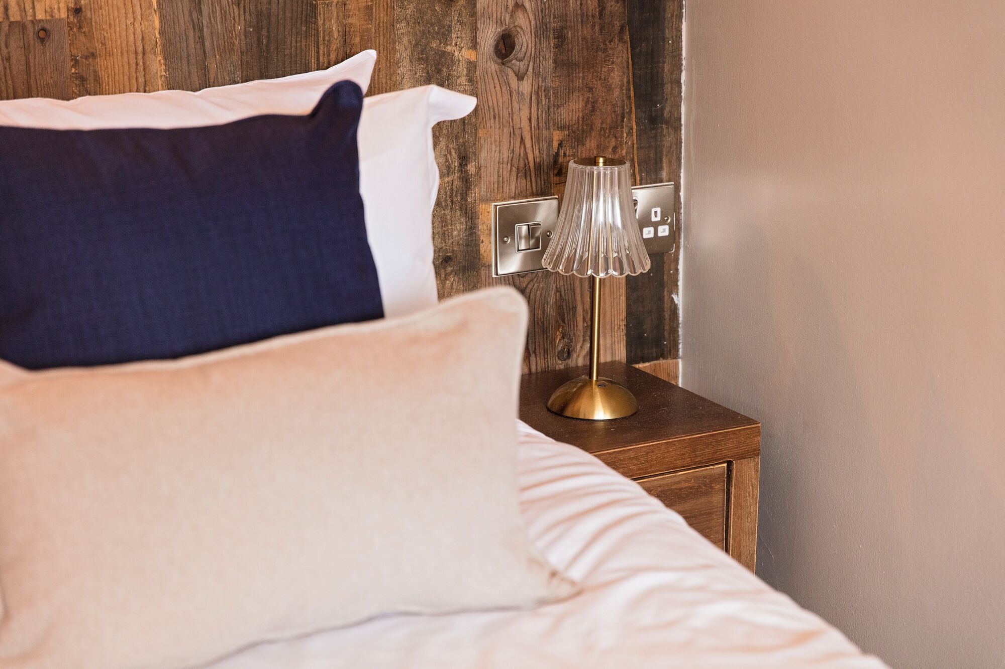 A bed with a white and navy pillow, a gold lamp on a wooden nightstand, and a switchboard on the wall.