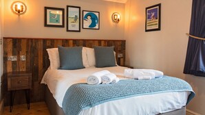 Una cama bien hecha con una manta azul y almohadas, cabecera de madera y un cuadro colgado en la pared.