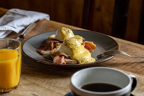 Un plato de desayuno con huevos Benedict, tocino y un vaso de jugo de naranja sobre una mesa de madera.