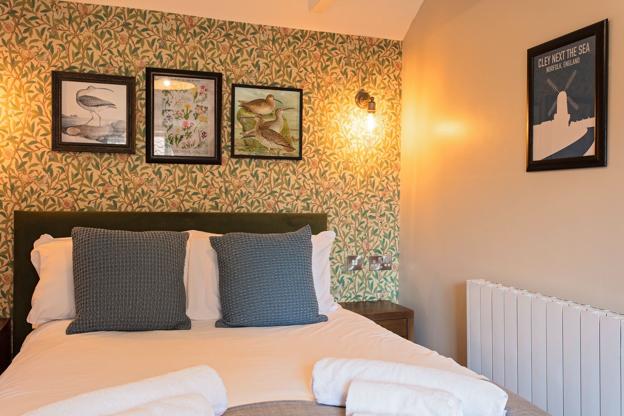 A neatly made bed with white and gray pillows, a framed botanical print, and a framed picture of birds on the wall.