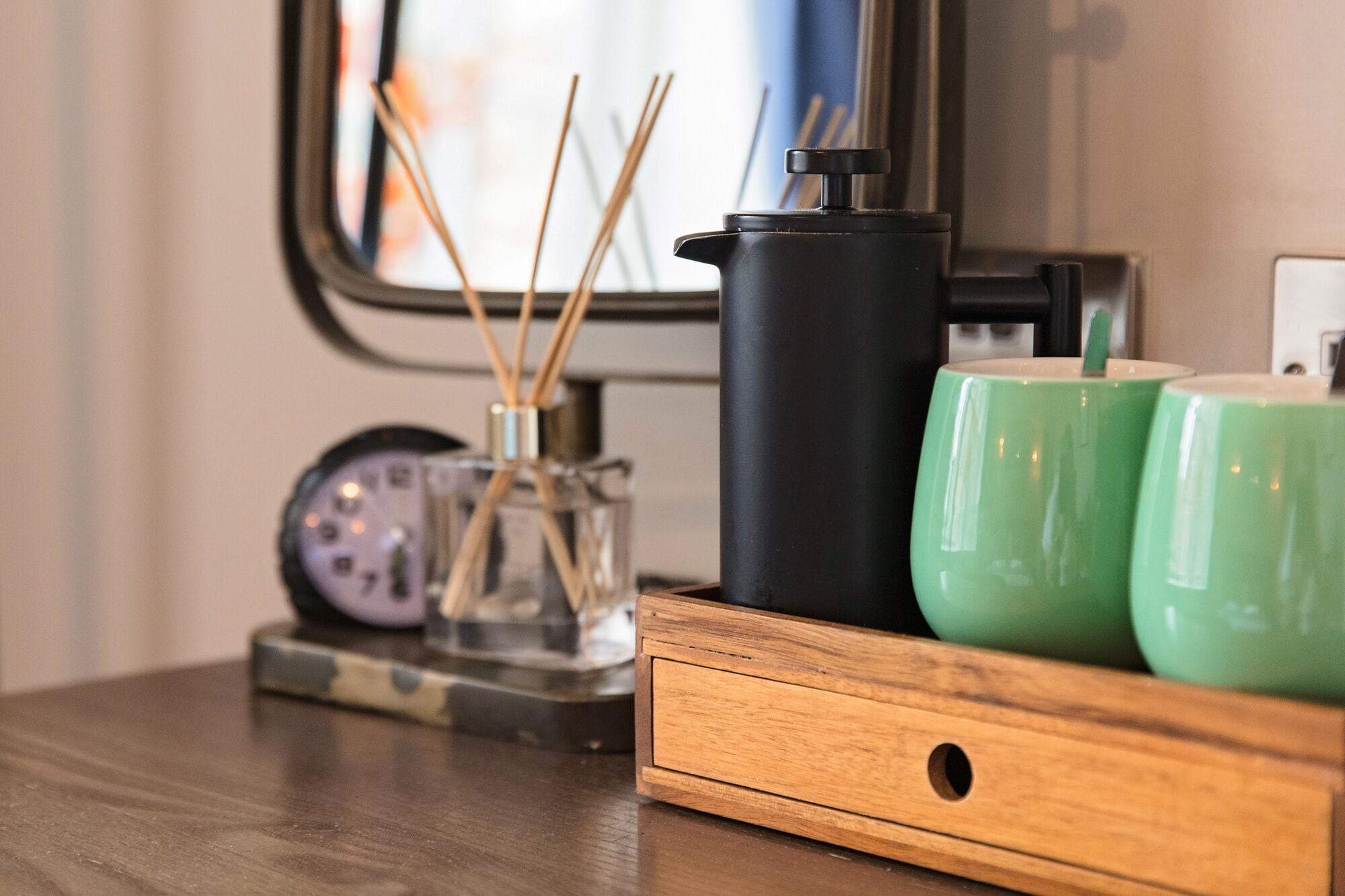 A wooden tray with a black coffee grinder, two green mugs, and a glass container with reed sticks.