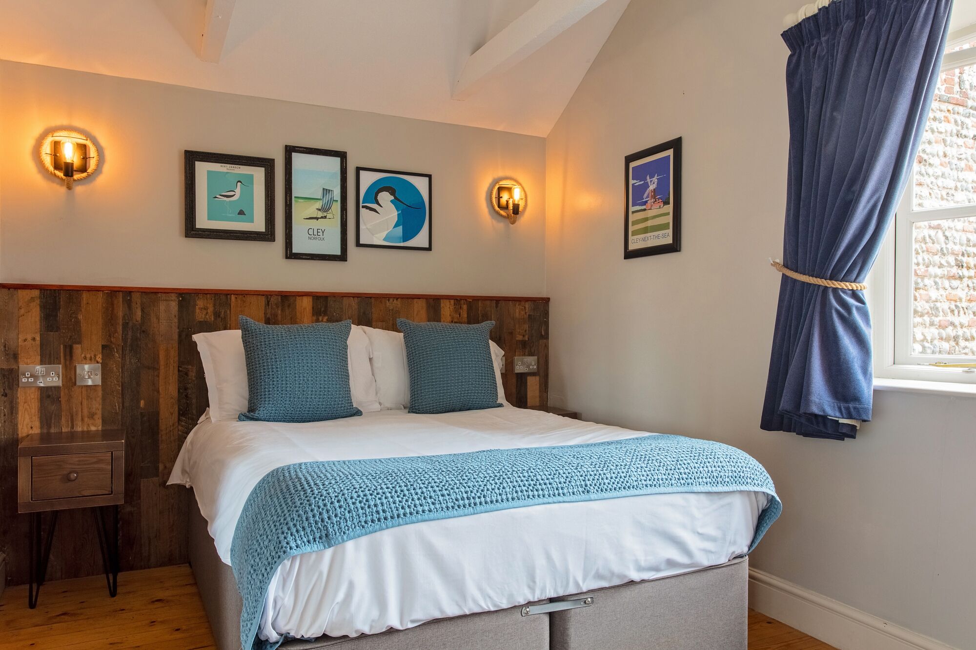 A bedroom with a wooden headboard, a bed with white and blue bedding, a nightstand, and wall-mounted lights.