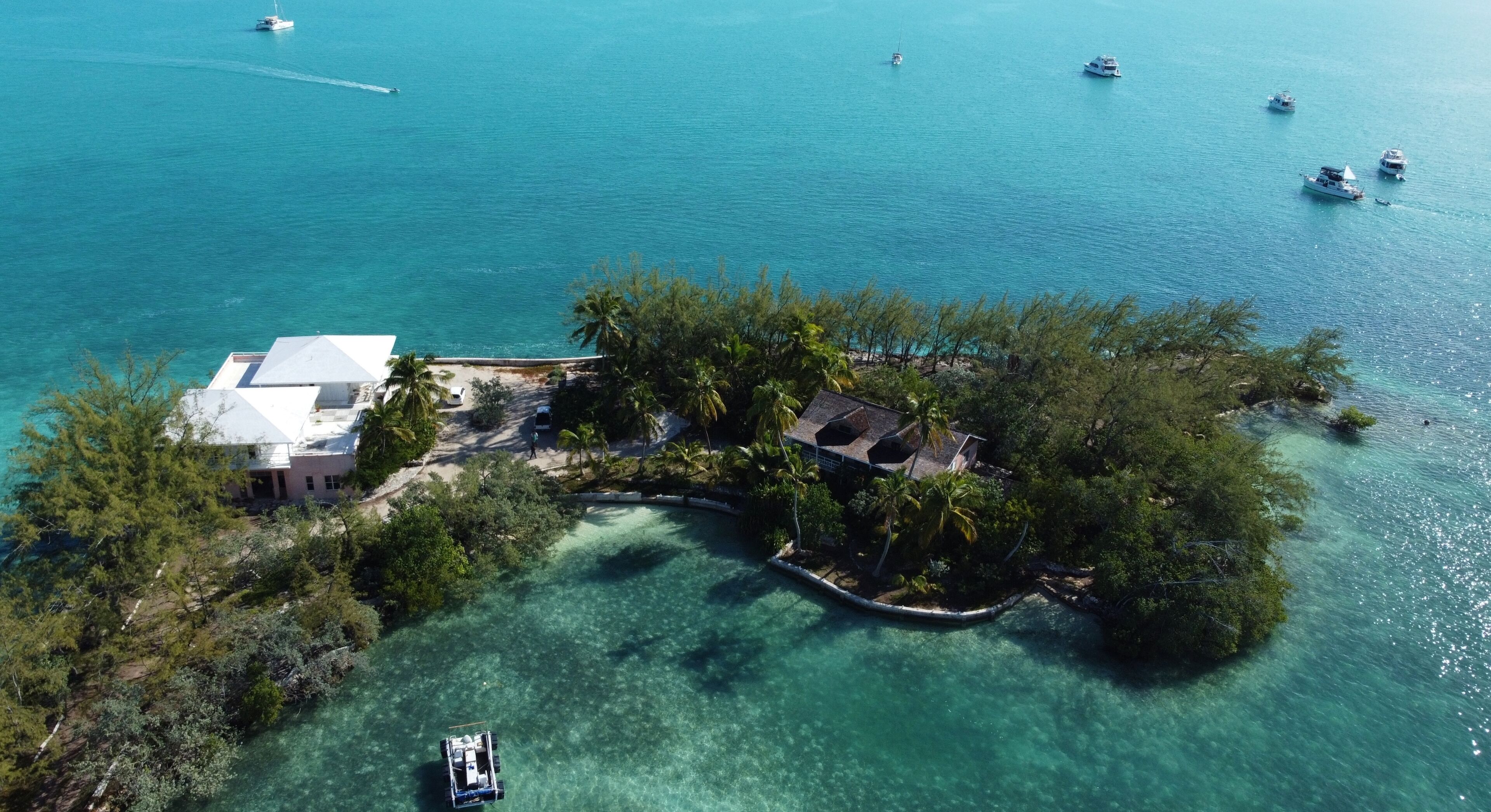 Vista aerea di un'isola tropicale con acque turchesi cristalline, una piccola casa con tetto bianco e una barca ormeggiata vicino alla riva.