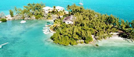 An aerial view of a tropical island with clear turquoise waters, surrounded by lush greenery and several white buildings, likely a resort or private property.