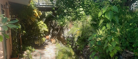 A narrow stone pathway flanked by lush greenery and potted plants, leading to a covered area with a wooden roof.