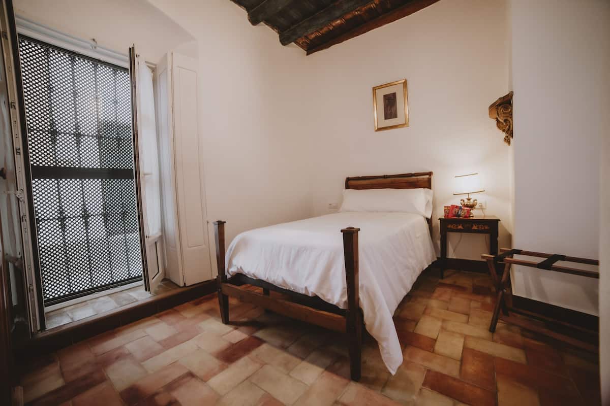A bedroom with a bed, a nightstand, a chair, a window with a metal screen, and a wooden ceiling.