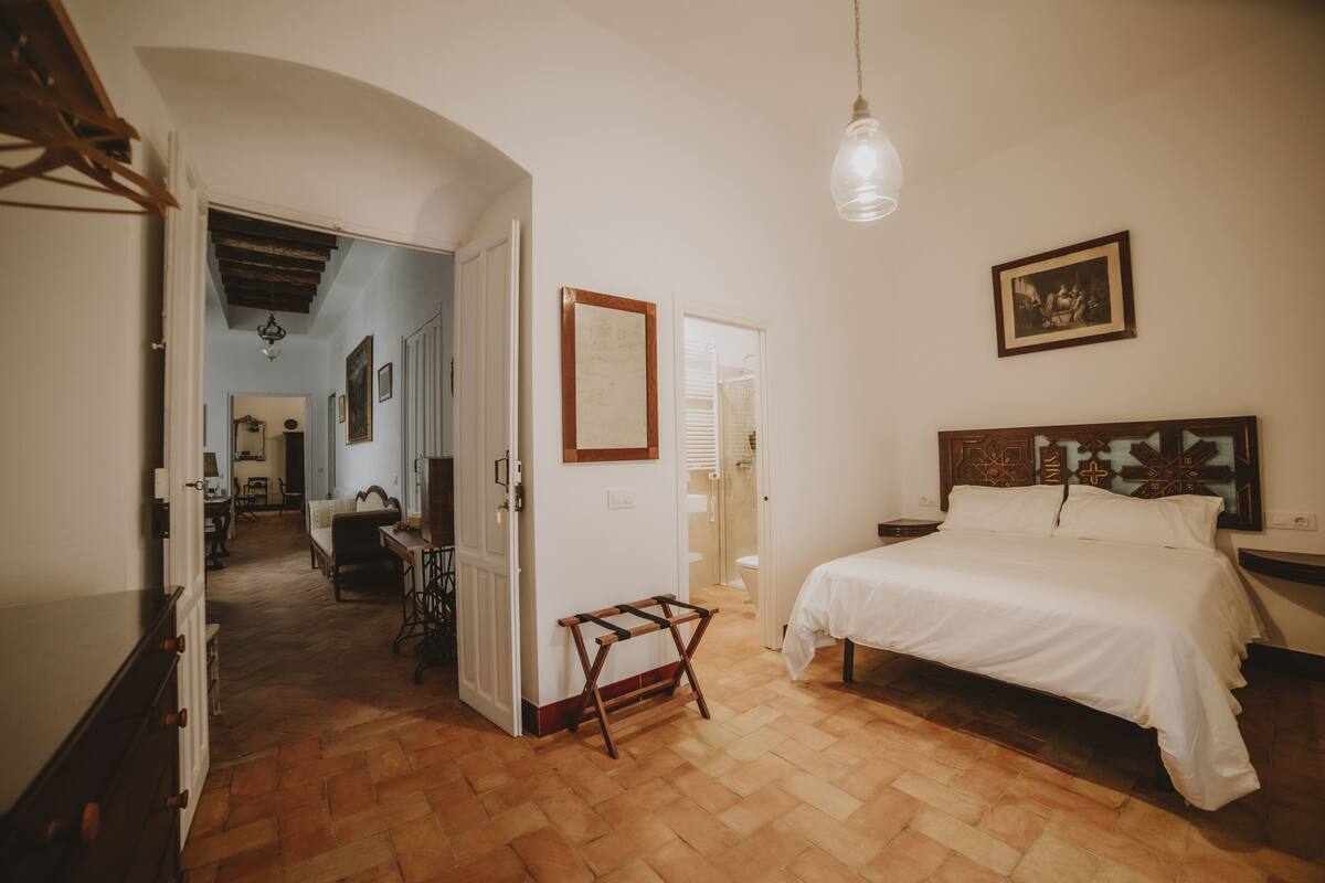 A bedroom with a bed, a wooden bench, a hanging light, and a door leading to another room.