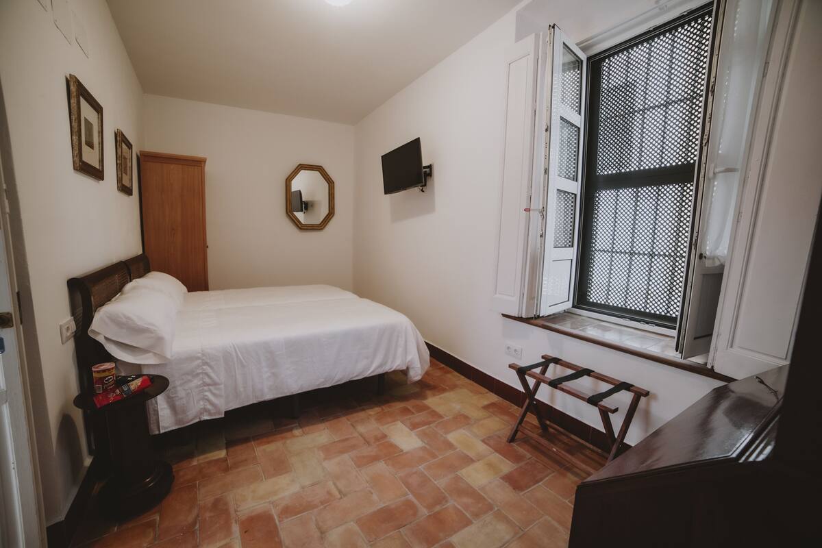 A small, clean room with a single bed, a wooden headboard, a small table with a chair, a window with a metal screen, and a door.