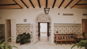 Un interior con suelo de baldosas, un banco de madera, plantas en macetas y un arco que da a otra habitación.