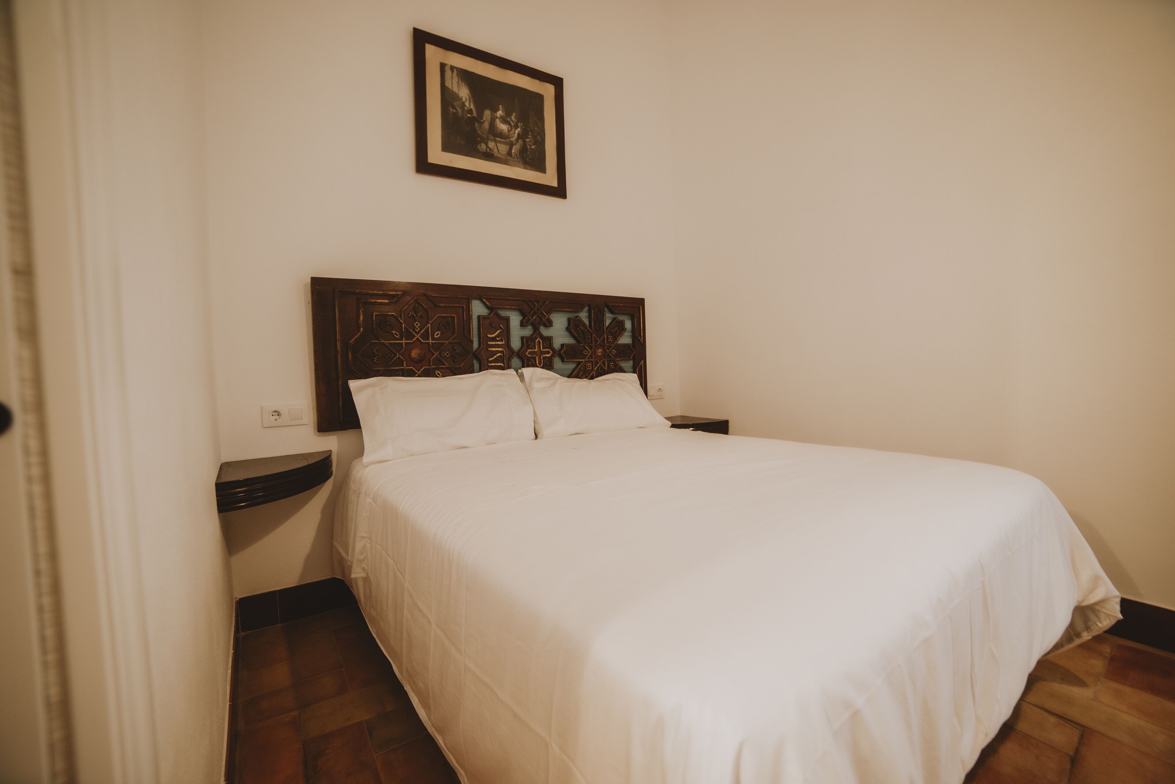 Un letto rifatto con cura, una testiera in legno, un comodino e un quadro incorniciato al muro.