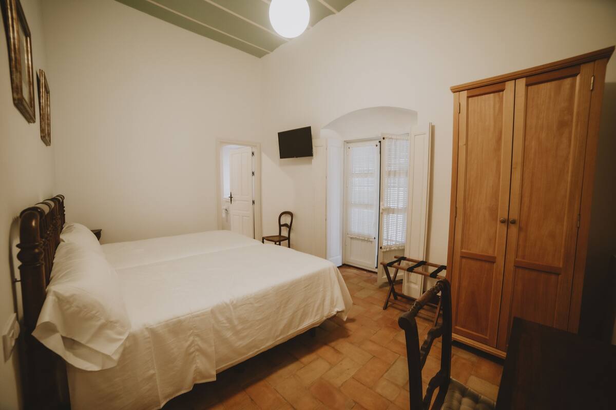 A bedroom with a single bed, a wooden wardrobe, a television mounted on the wall, and a door leading to another room.