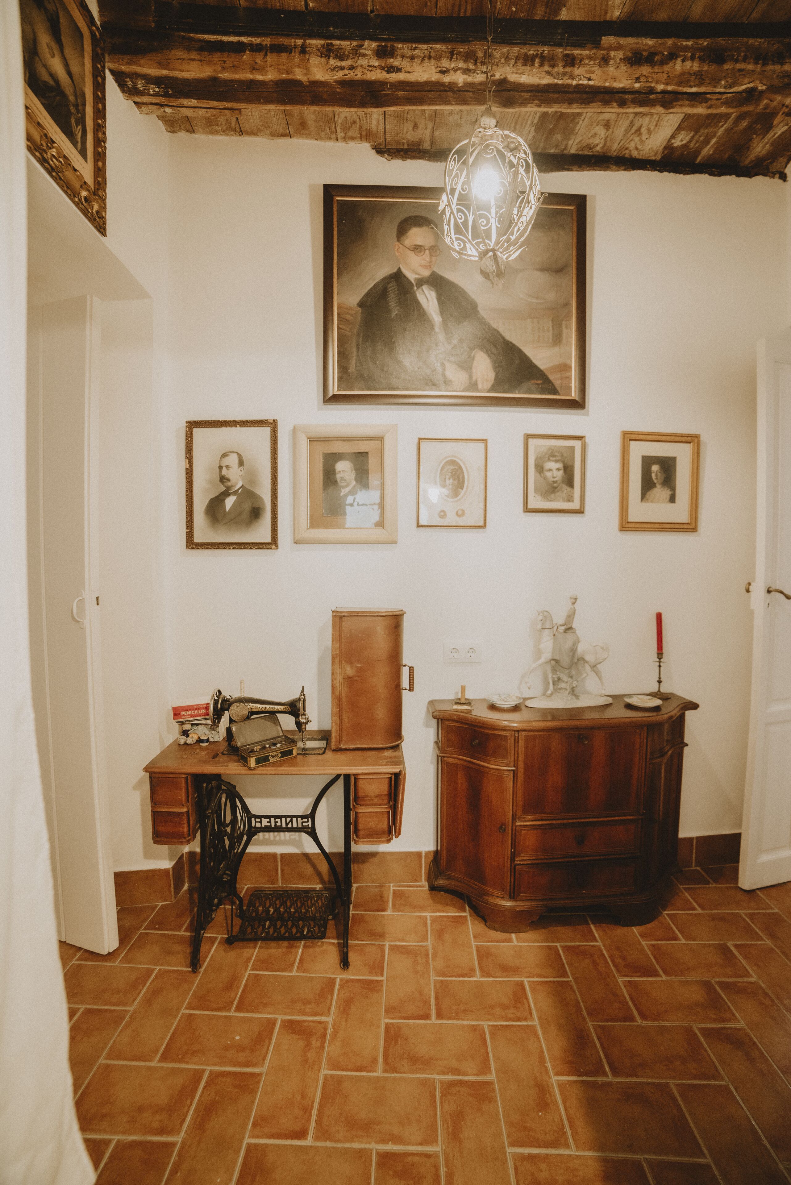 Een kamer met een houten bureau, een naaimachine, een houten kast en een wand versierd met ingelijste portretten.