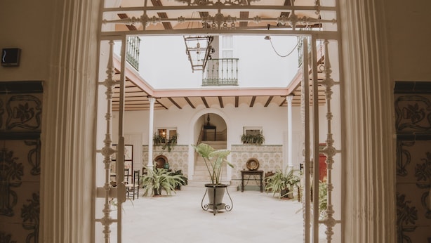 Un arco de entrada da a un patio con suelo de baldosas, plantas en macetas y un balcón con barandilla.