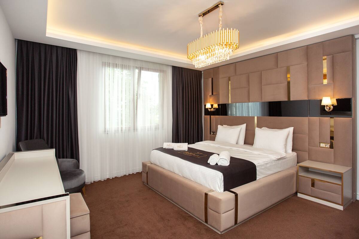 Una habitación de hotel moderna con una cama grande, un escritorio y una silla. Hay una araña colgante sobre la cama y lámparas de pared a cada lado.