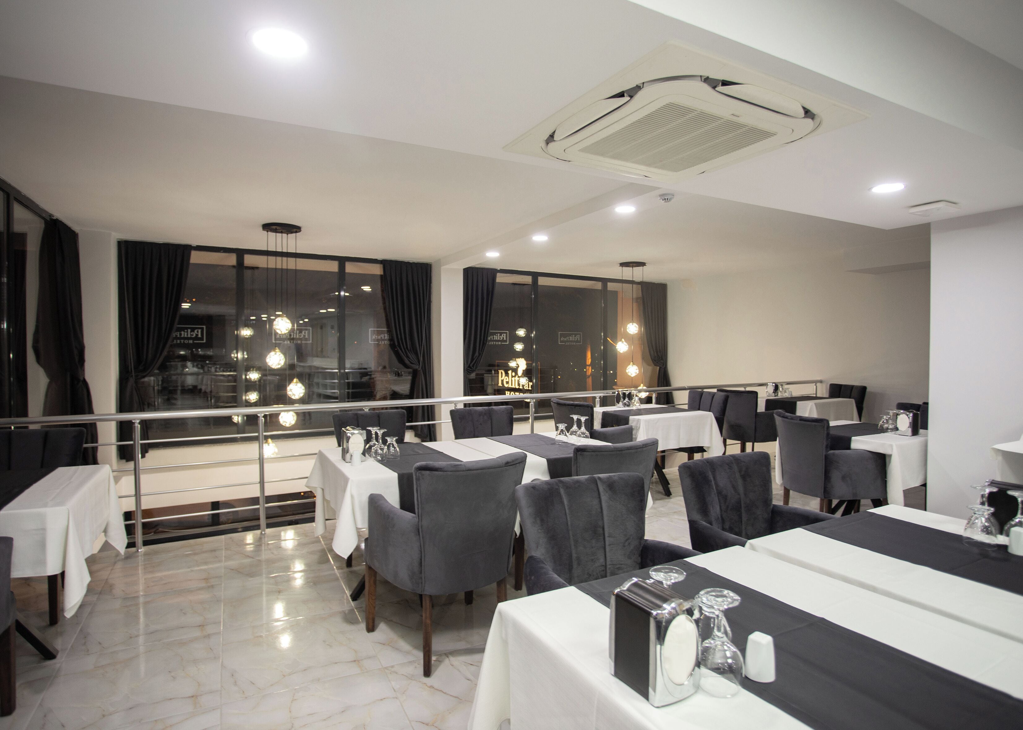 A restaurant with tables set for dining, a balcony with a railing, and large windows with curtains.