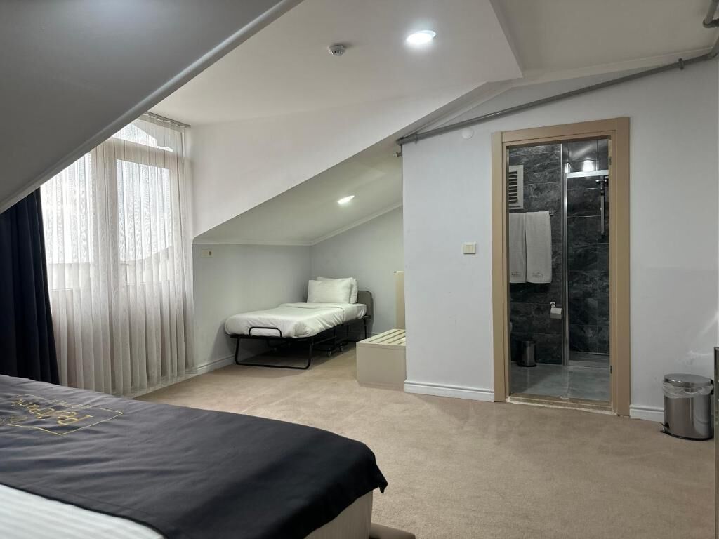 A bedroom with a bed, a small desk, a chair, a window with curtains, and a bathroom with a glass door.