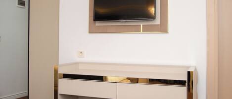A modern, minimalist room with a white desk, a flat-screen TV mounted above it, and a cushioned bench at the desk's base.