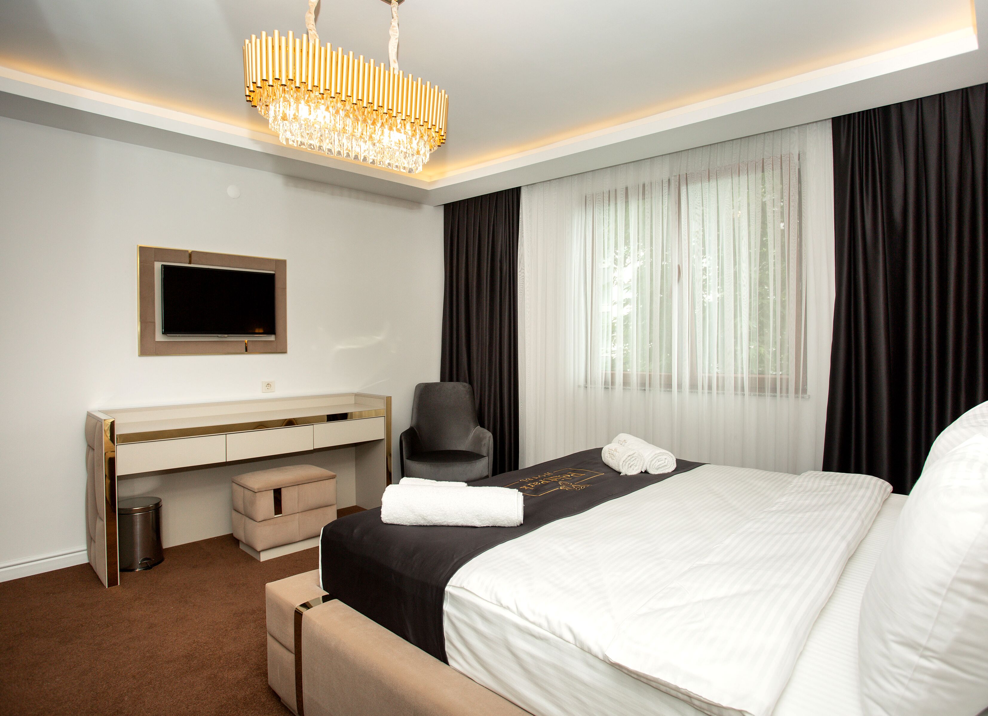 A modern hotel room with a large bed, a desk, a chair, a TV, and a chandelier.