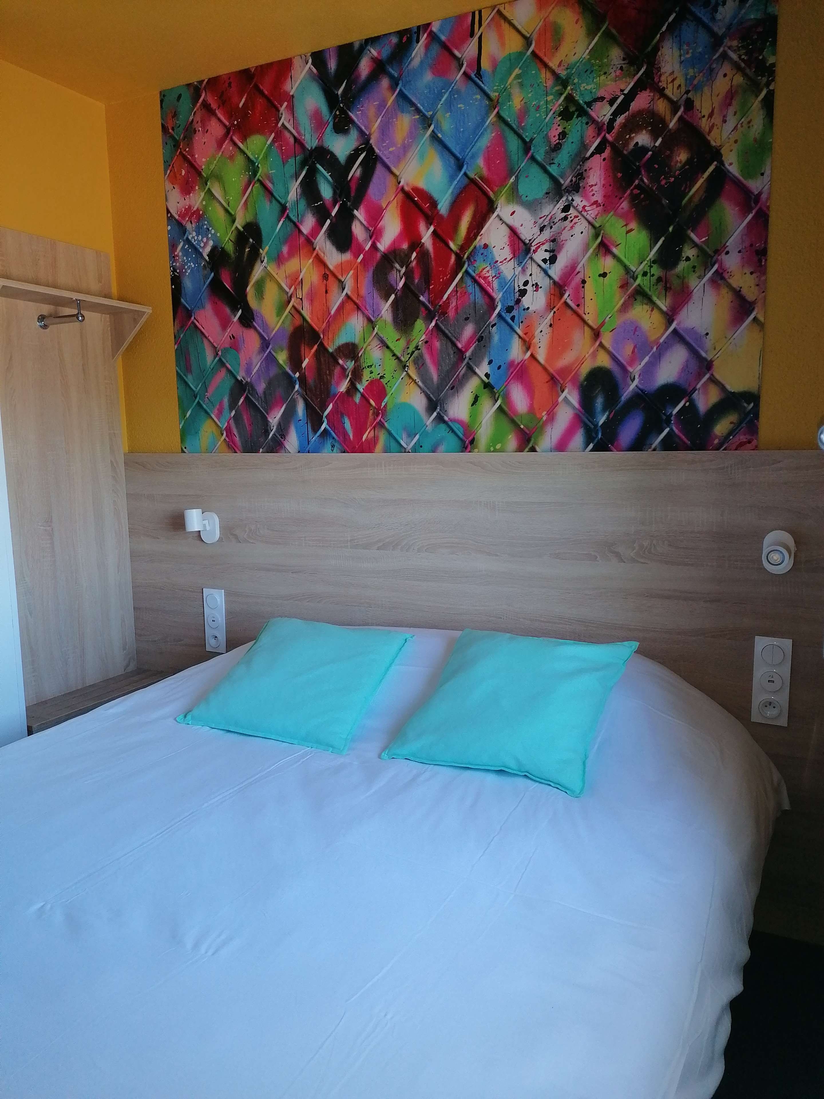 Une chambre d’hôtel avec un lit, deux oreillers, une tête de lit en bois, une peinture abstraite aux couleurs vives accrochée au mur et un interrupteur.