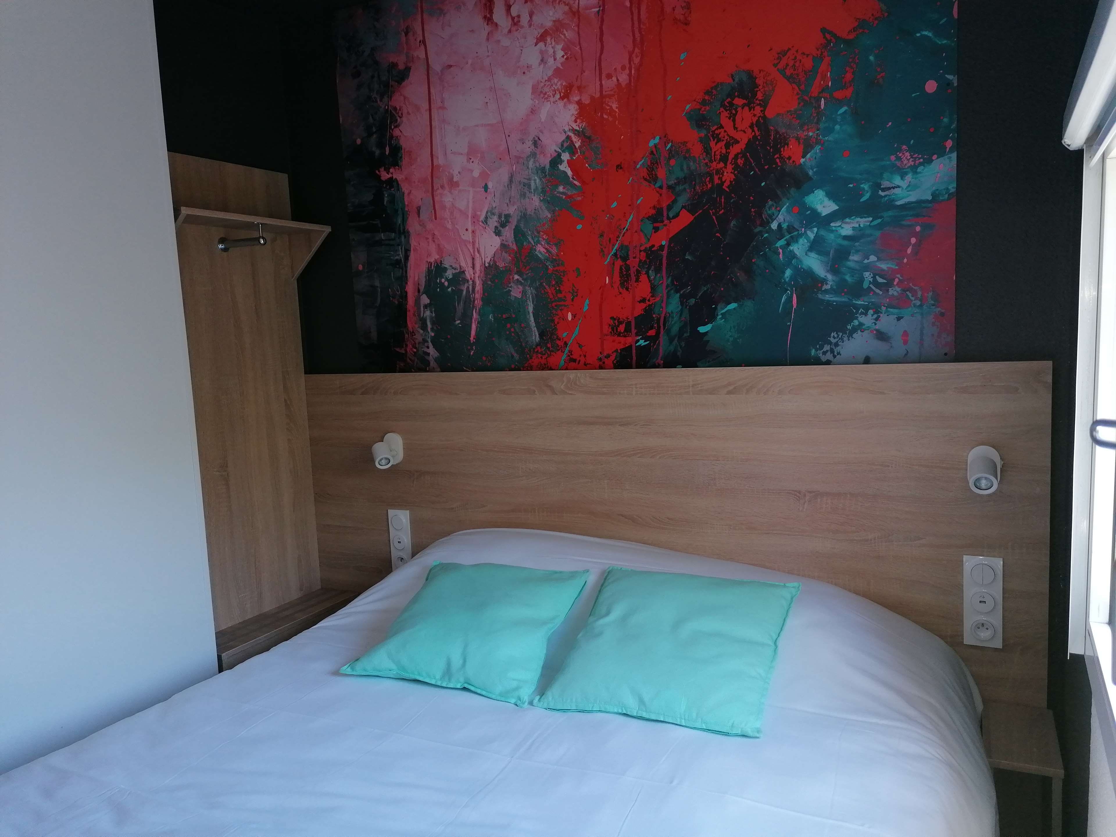 Una cama individual con colchón blanco y dos almohadas verdes, cabecera de madera y una pared con un cuadro abstracto de colores.